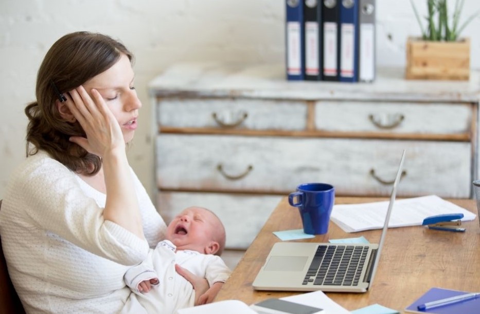 Postpartum depressed mom crying with baby in her lap sitting at a desk trying to work